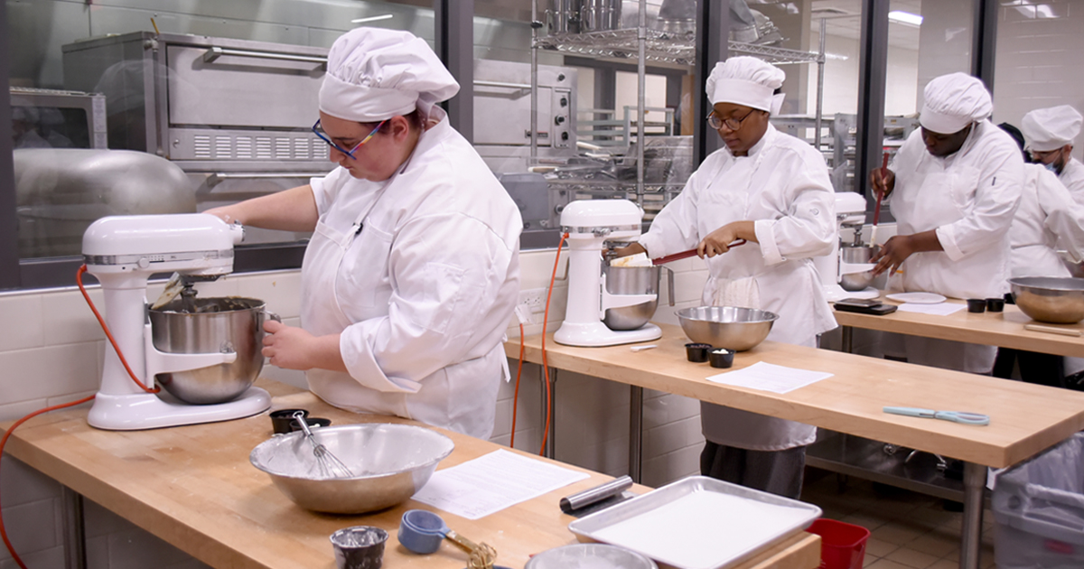 People in chef uniforms using standing mixers in the campus kitchen lab