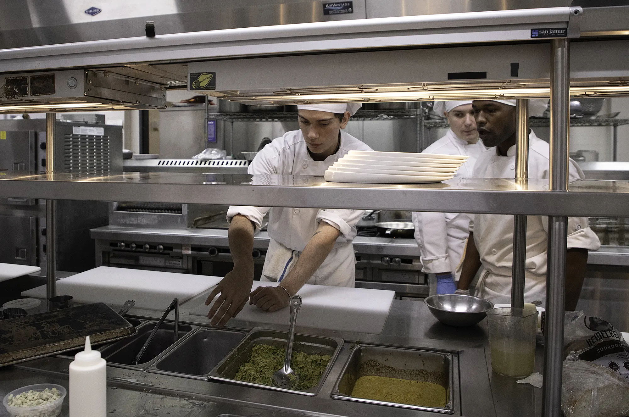 Culinary arts students in white chef coats and hats prepare food at a stainless-steel service line, arranging ingredients and plates in a commercial teaching kitchen