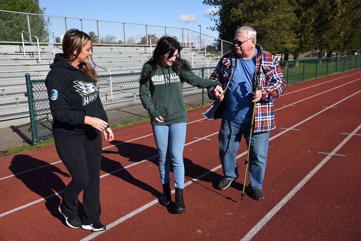 Student helping man with cane walk on track with professor supervision