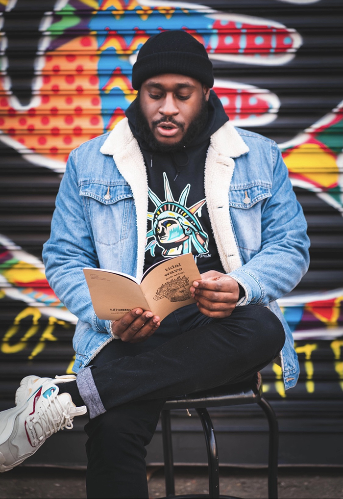 Image of Kofi Antwi reading from his book of poetry, sitting in front of colorful painted wall