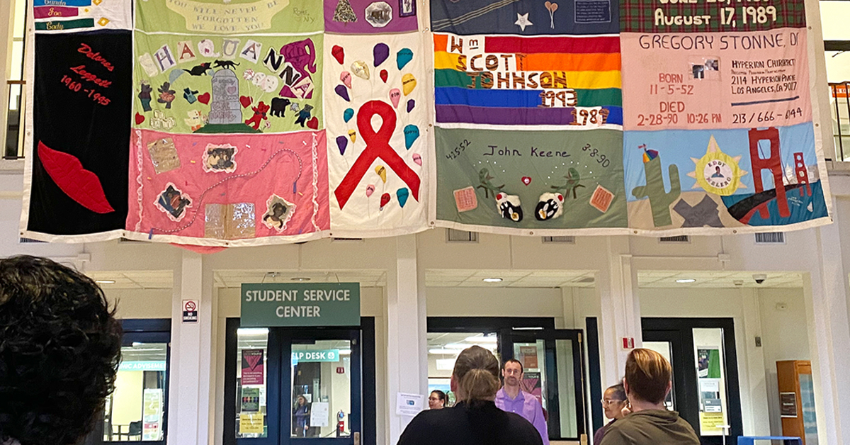 Image shows the people standing looking up at a display of AIDS Memorial Quilt panels at MVCC